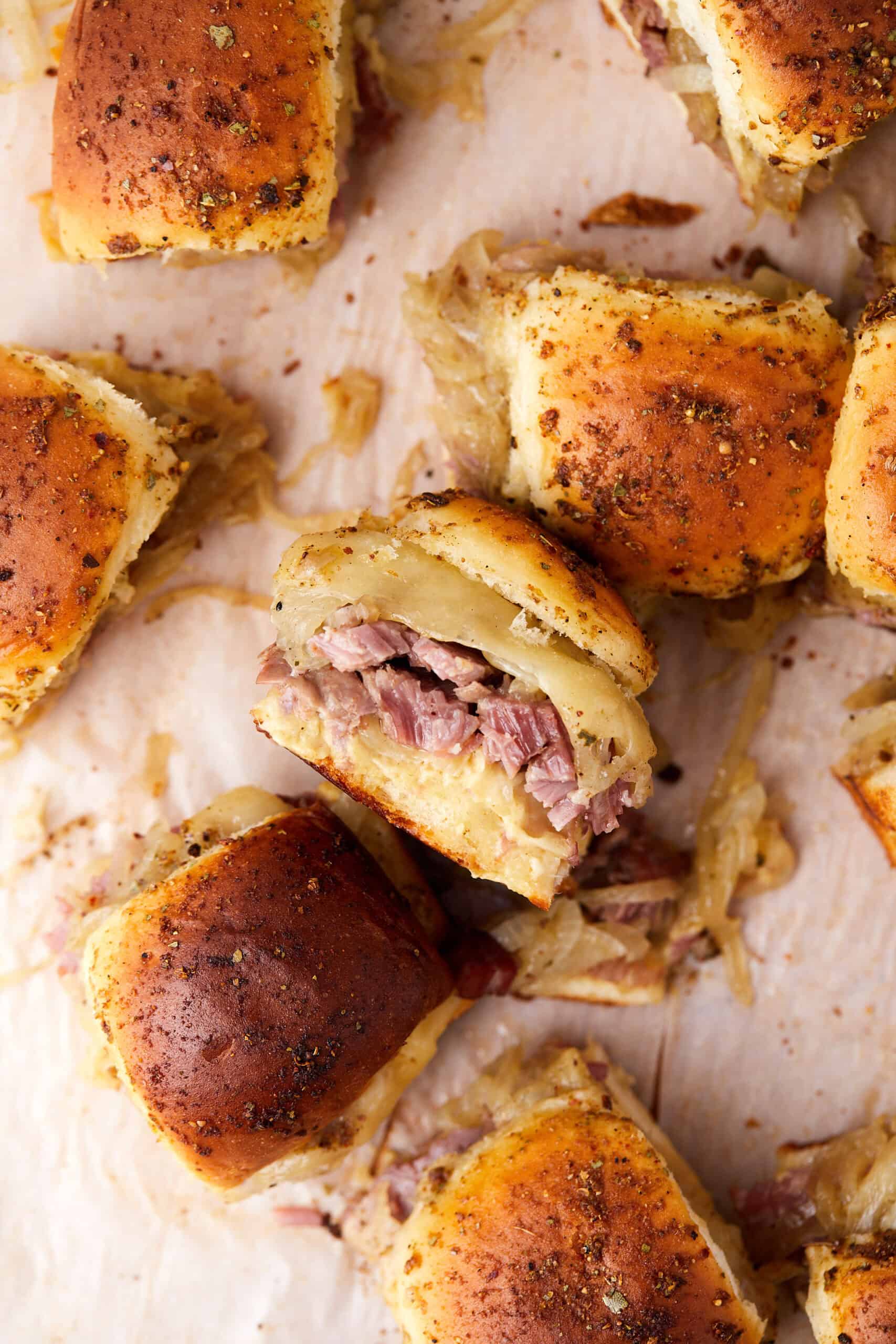 A close-up of mini sandwiches on golden-brown rolls, filled with shredded meat and melted cheese, arranged on parchment paper with herbs and caramelized onions. One sandwich in the center is cut open to show the filling.