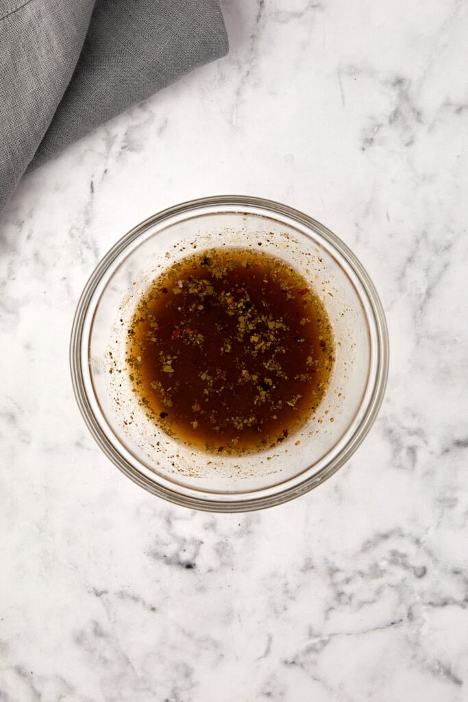 A glass bowl filled with brown liquid and herbs sits on a white marble surface, with part of a gray cloth visible in the top left corner.