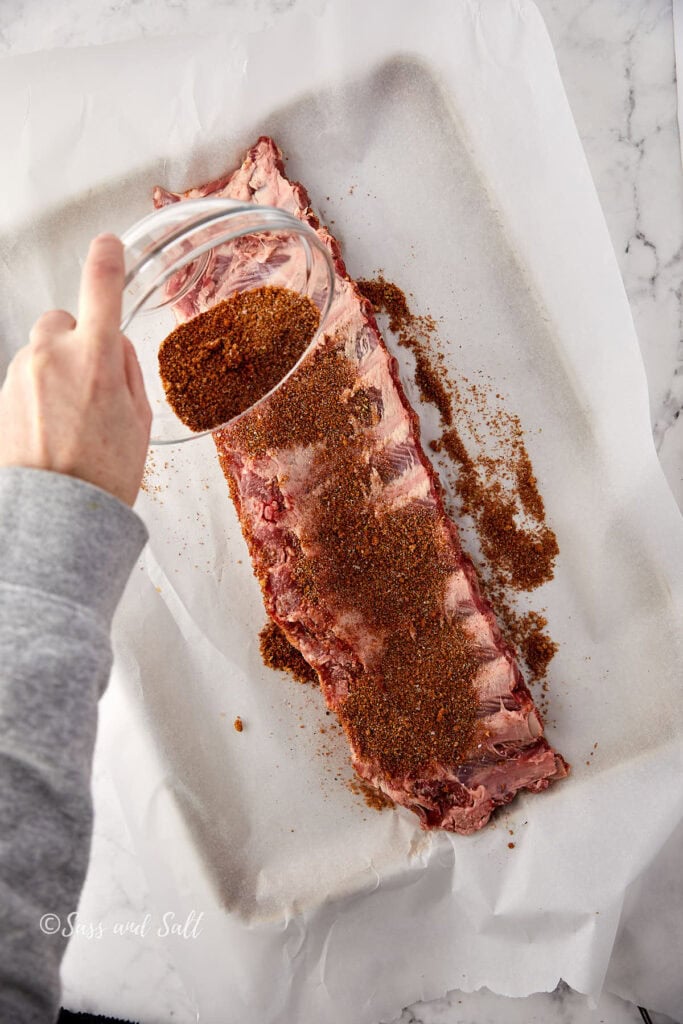A person sprinkles a spice rub onto a rack of raw ribs placed on parchment paper, preparing them for cooking. The image is taken from above, showing the ribs on a marble countertop.