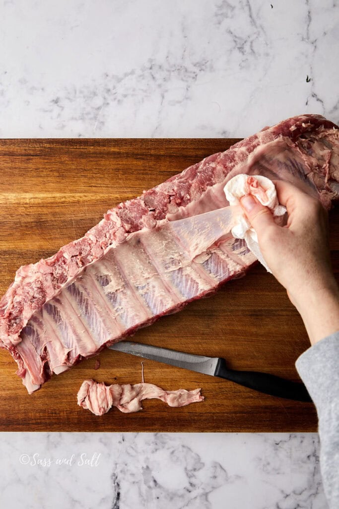 A person removes the membrane from a rack of raw ribs on a wooden cutting board. A knife rests nearby, and the scene is set on a marble countertop.