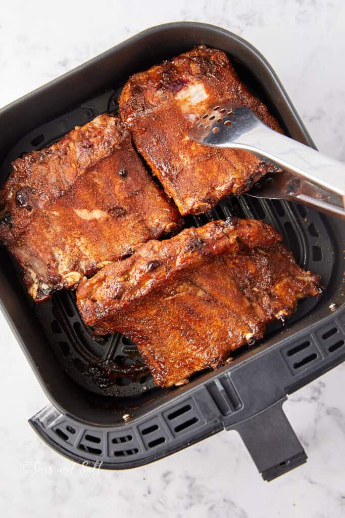Four pieces of seasoned ribs sit in an air fryer basket, with a pair of metal tongs resting on top. The ribs are cooked to a dark brown color, suggesting they are well-seasoned and roasted. The background is a white marble surface.