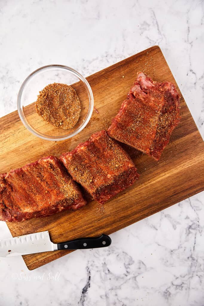A wooden cutting board with three racks of ribs seasoned with spices. A clear bowl filled with more spice mix is to the side. A chef's knife rests on the board. The backdrop is a white marble surface.