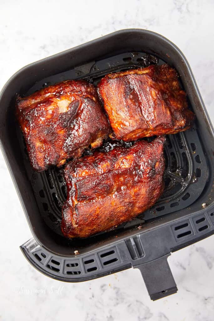 Three pieces of glazed, crispy pork ribs in an air fryer basket. The ribs are coated with a rich, dark sauce, glistening under the light, set on a light marbled countertop.