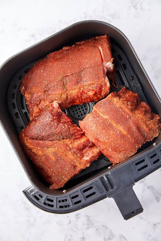 Raw, seasoned ribs arranged inside an air fryer basket. The ribs are coated with a dark spice rub on a marble-like countertop.
