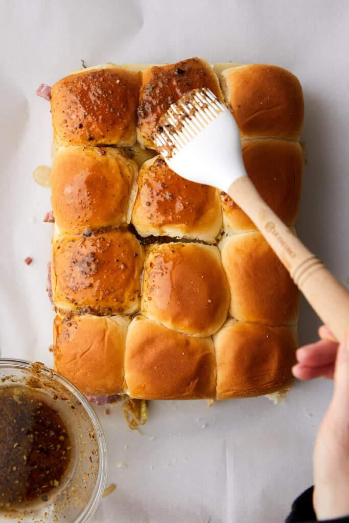 A hand uses a pastry brush to spread a seasoning mixture over a batch of baked slider rolls. A small bowl with seasoning is nearby on the white surface.