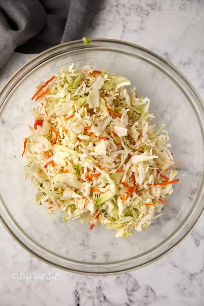 A clear glass bowl filled with shredded cabbage, carrots, and other vegetables sits on a marble surface. A gray cloth is partially visible in the top left corner.