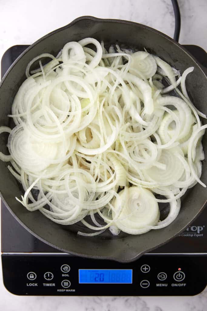 A skillet filled with thinly sliced onions sits on an electric cooktop with a blue digital timer display, preparing to be cooked.