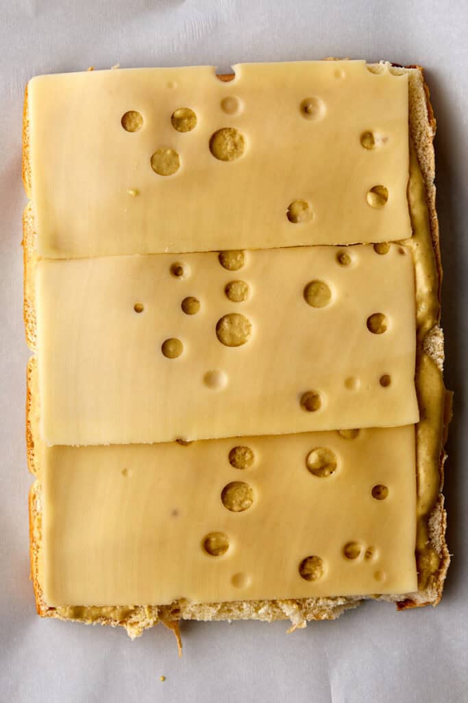 Three slices of Swiss cheese with holes are laid on top of a piece of bread that is spread with horseradhish mustard sauce viewed from above on a sheet of parchment paper.