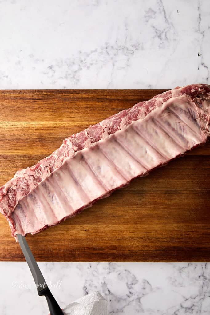 Raw pork ribs on a wooden cutting board, placed on a marble surface. A knife is partially visible on the left side of the image.