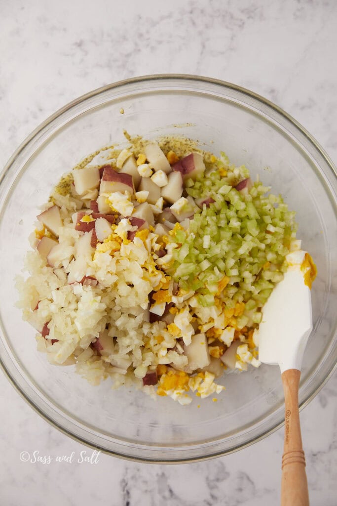 A glass bowl contains chopped red potatoes, celery, onion, eggs, and seasonings, ready to be mixed with a white spatula on a marble surface.