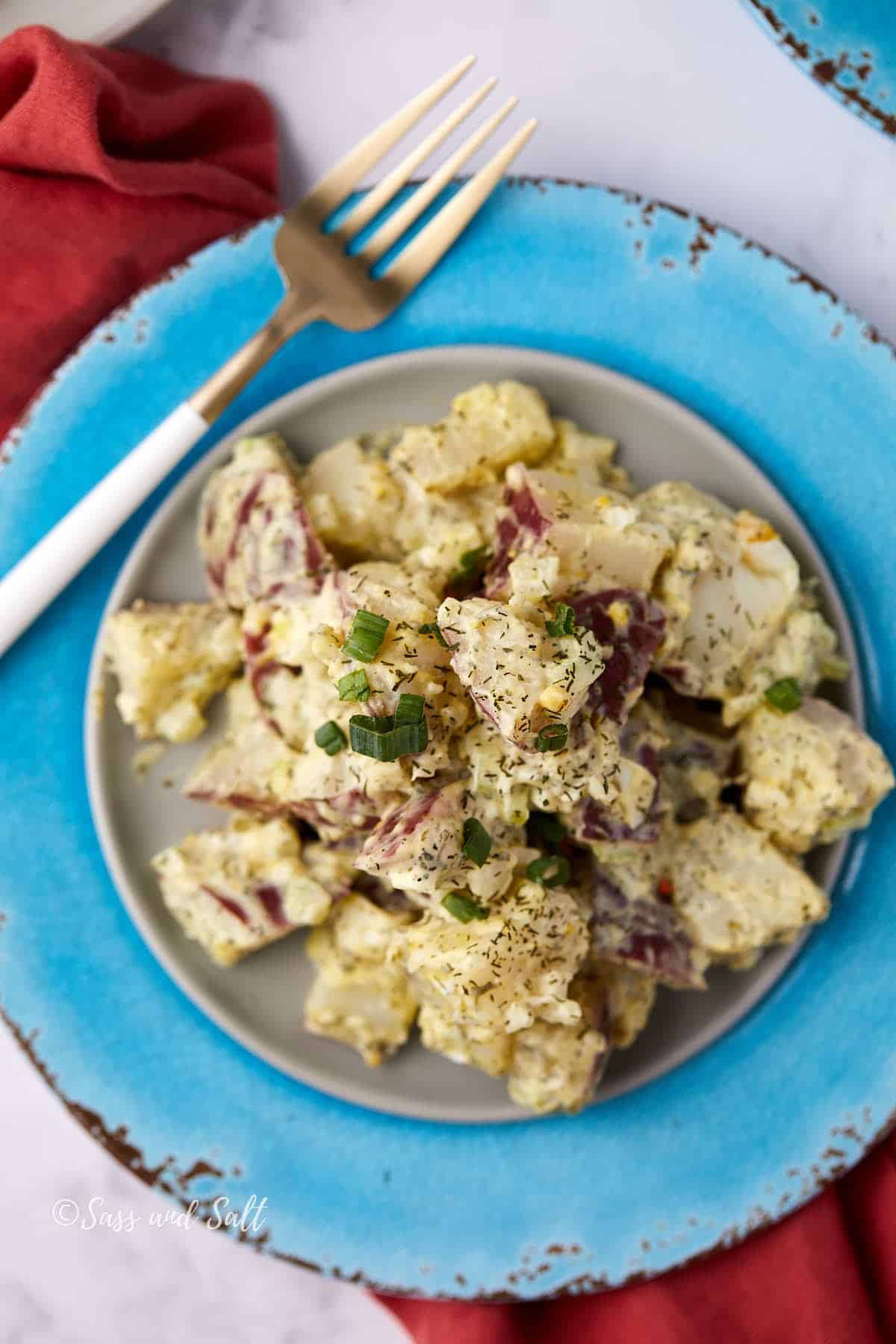 A plate of creamy potato salad with chopped green onions on top, served on a light gray plate with a distressed blue charger and a gold and white fork nearby.