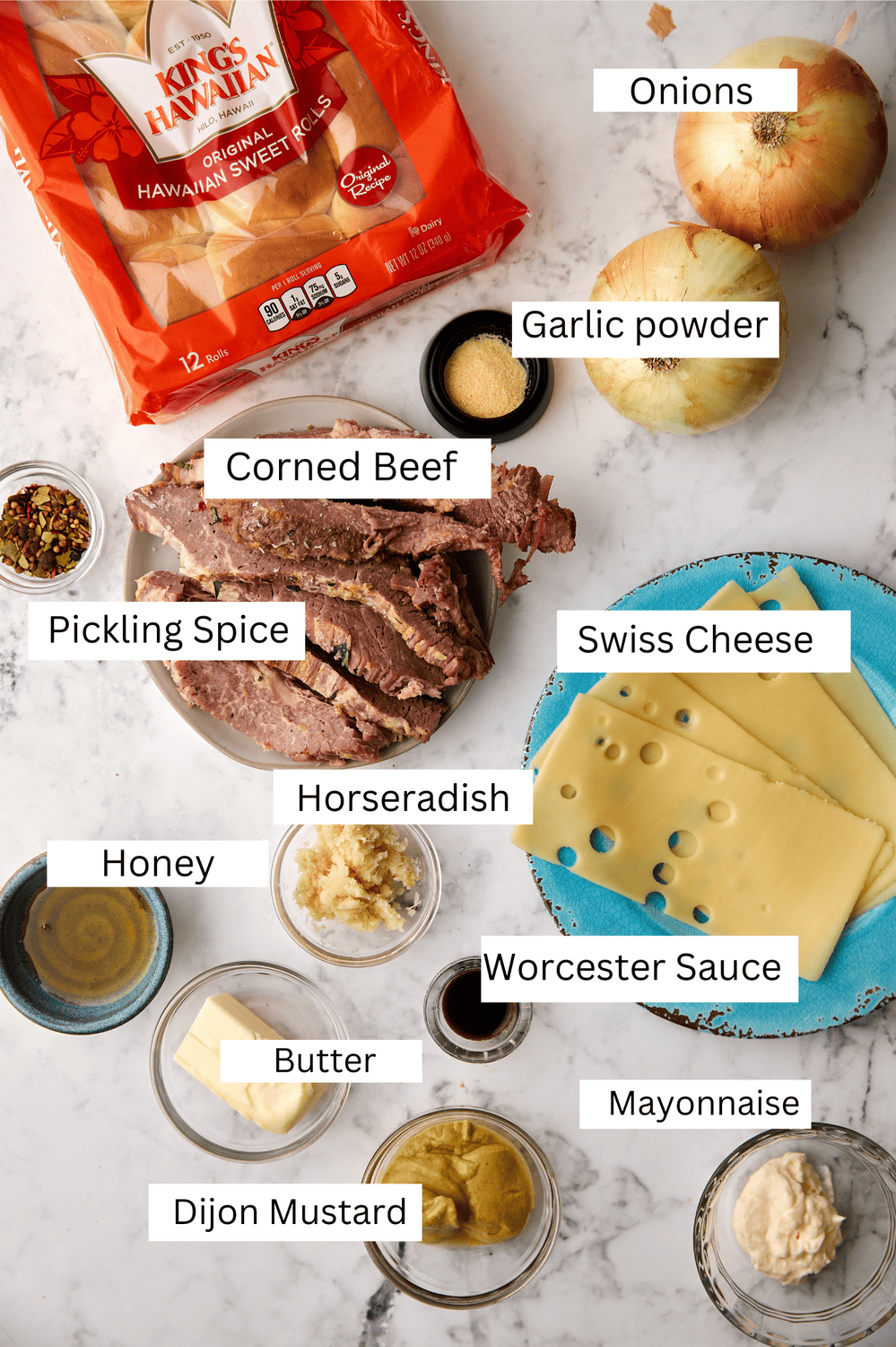 Top-down view of labeled ingredients for a corned beef sandwich, including Hawaiian sweet rolls, corned beef, Swiss cheese, onions, garlic powder, pickling spice, horseradish, honey, Worcestershire sauce, butter, Dijon mustard, and mayonnaise.