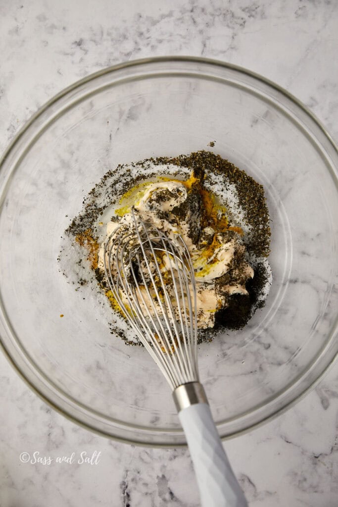 A glass bowl on a marble countertop contains butter, dried herbs, spices, and oil being mixed with a metal whisk.