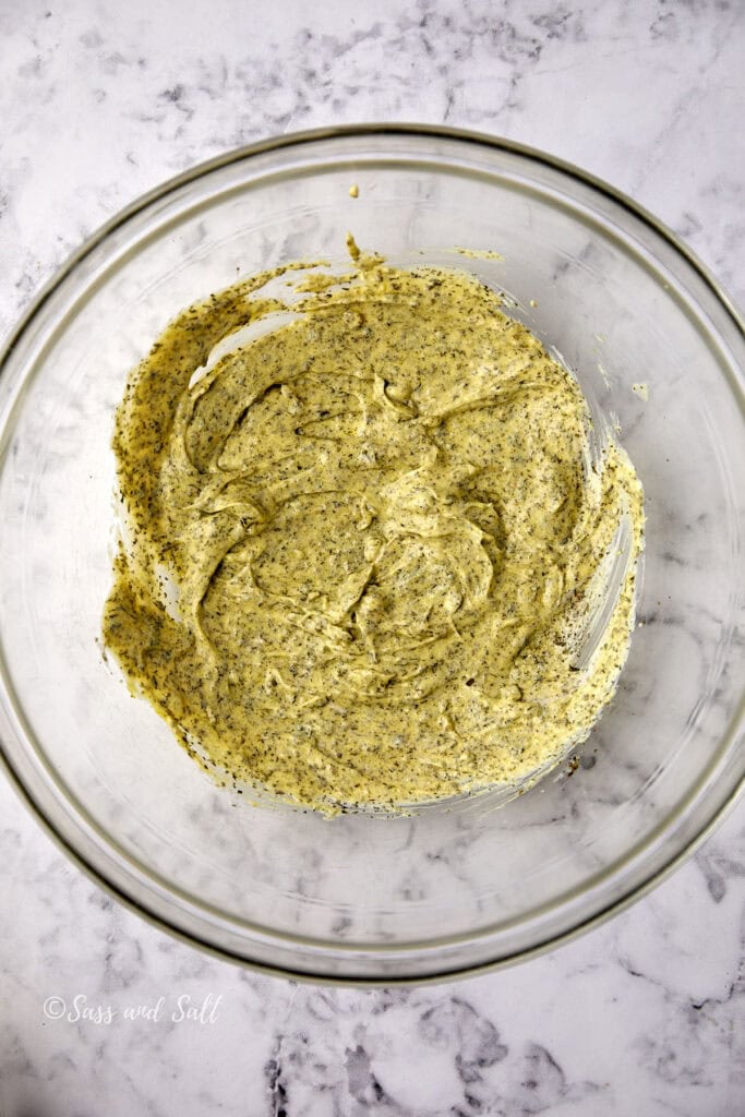 A glass mixing bowl with a creamy greenish-yellow herb and spice mixture, sitting on a white marble countertop.
