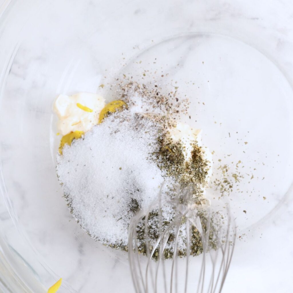 A metal whisk in a glass bowl with mayonnaise, sugar, dried herbs, black pepper.