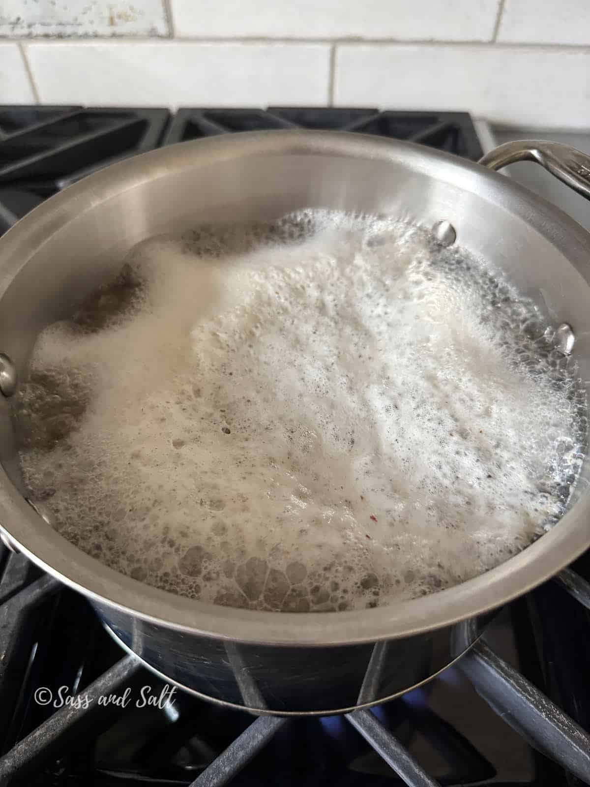 A stainless steel pot filled with bubbling foam sits on a gas stove, boiling over slightly. White subway tile backsplash and part of a metal handle are visible in the background.