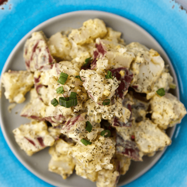 A plate of creamy potato salad with chunks of red-skinned potatoes, garnished with chopped green onions and herbs, served on a gray dish atop a blue placemat.