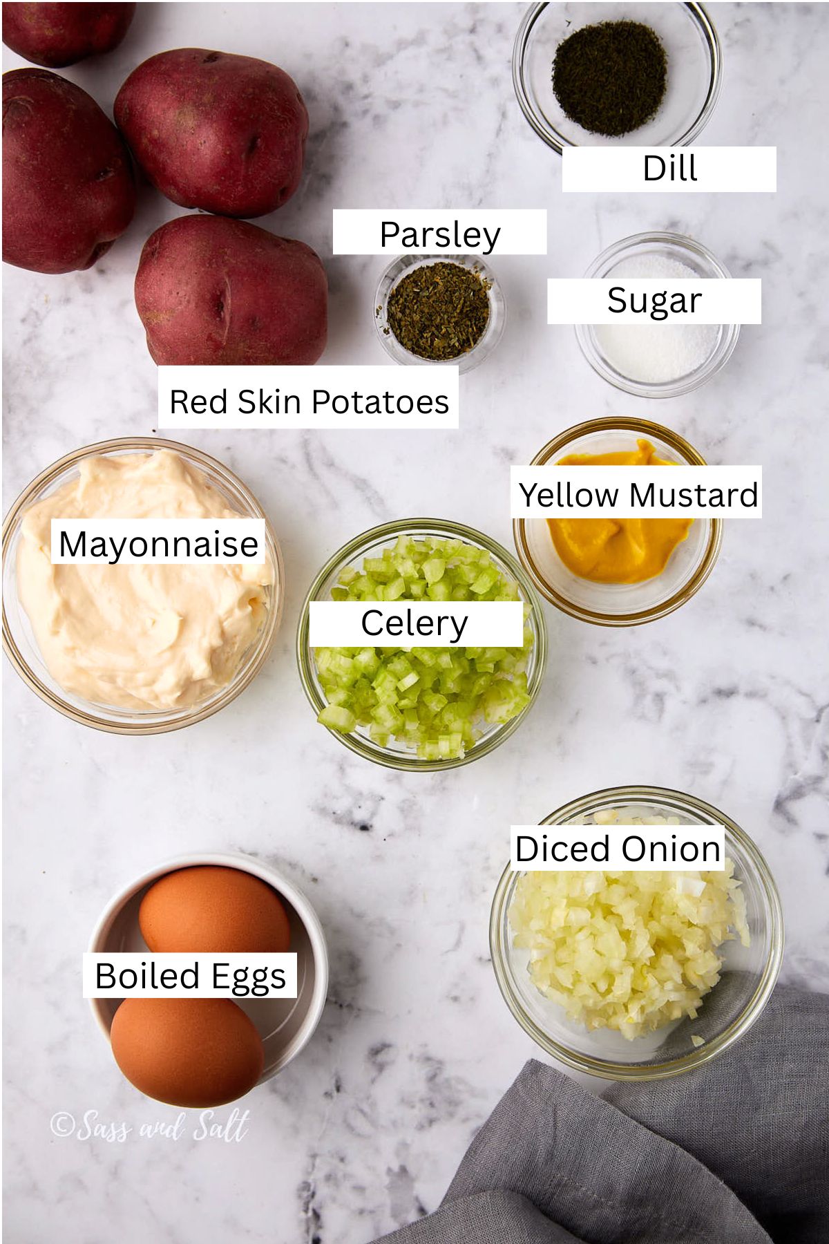 A flat lay of labeled ingredients for red skin dill potato salad on a marble surface: red skin potatoes, mayonnaise, boiled eggs, celery, diced onion, yellow mustard, dill, parsley, and sugar.