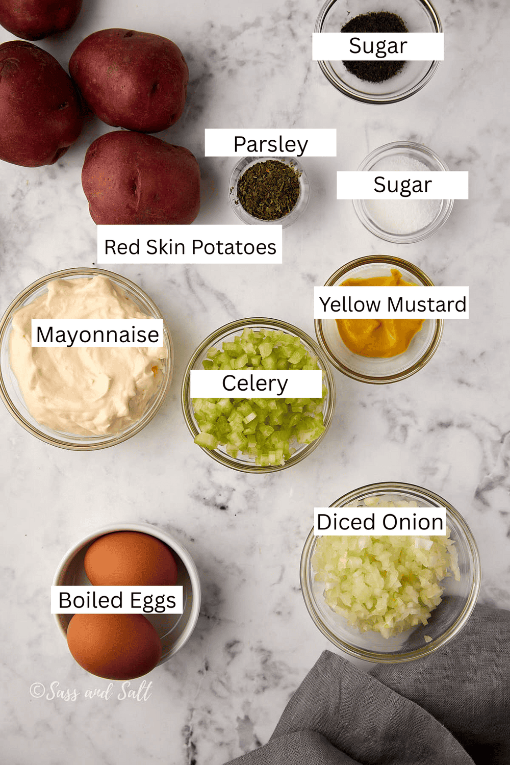Ingredients for potato salad arranged on a marble surface, including red skin potatoes, parsley, sugar, mayonnaise, celery, yellow mustard, diced onion, and boiled eggs, all in separate bowls or whole.
