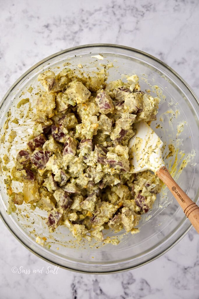 A glass bowl filled with creamy potato salad, mixed with herbs and diced red potatoes. A rubber spatula with a wooden handle rests inside the bowl on a marble countertop.