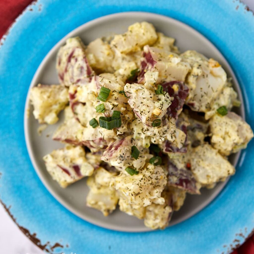 A plate of creamy red skin potato salad with chopped green onions and herbs, served on a gray plate with a blue rim.