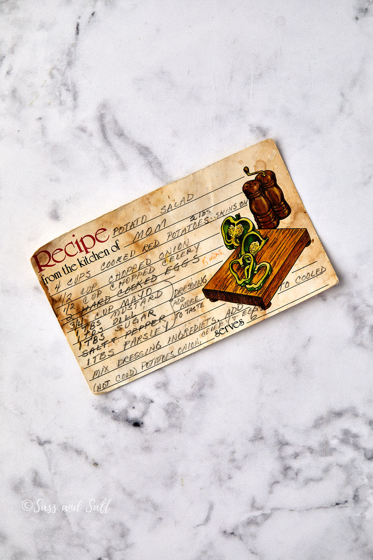 A vintage, handwritten recipe card for potato salad with a small illustration of a cutting board, knife, and vegetables, placed on a white marble surface. The card shows signs of age with stains and worn edges.