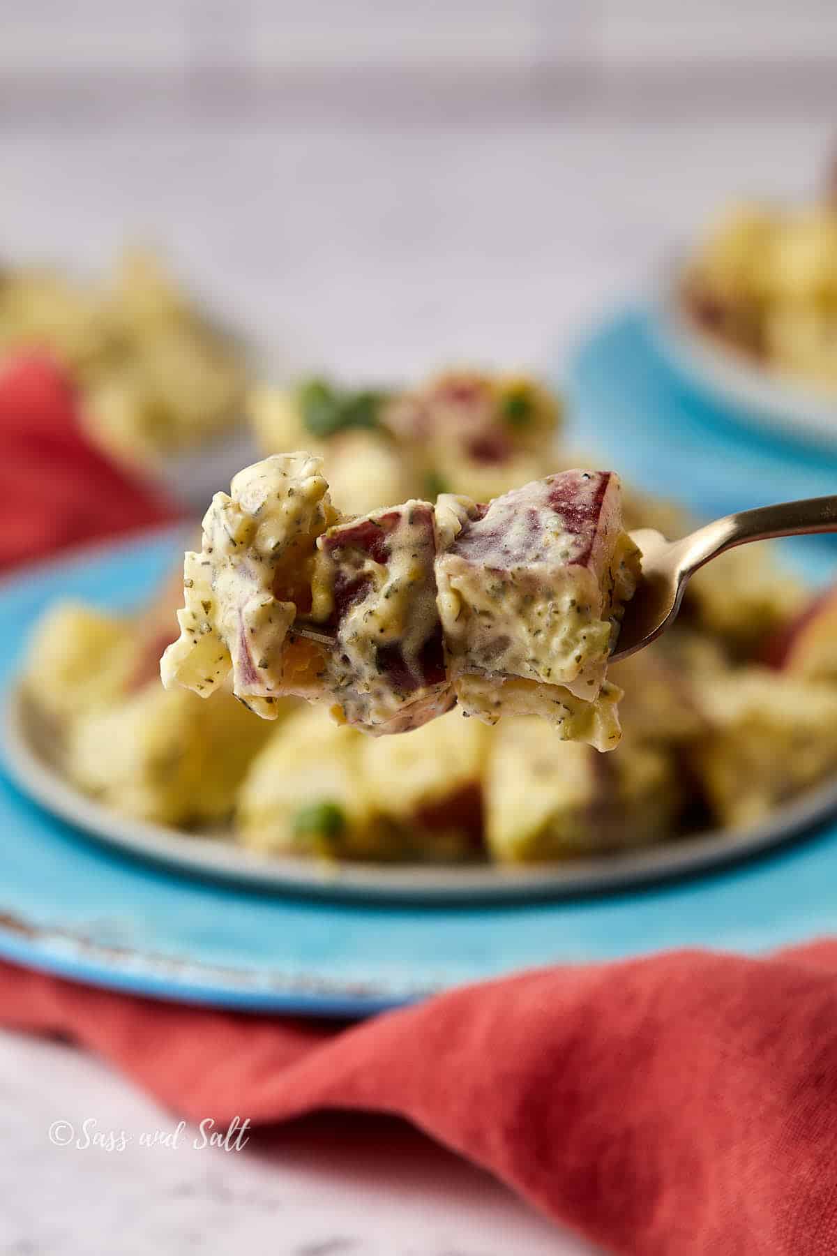 A fork holds up a bite of creamy potato salad with herbs, red potato skin visible. The background shows more potato salad on a blue plate and a red cloth napkin.