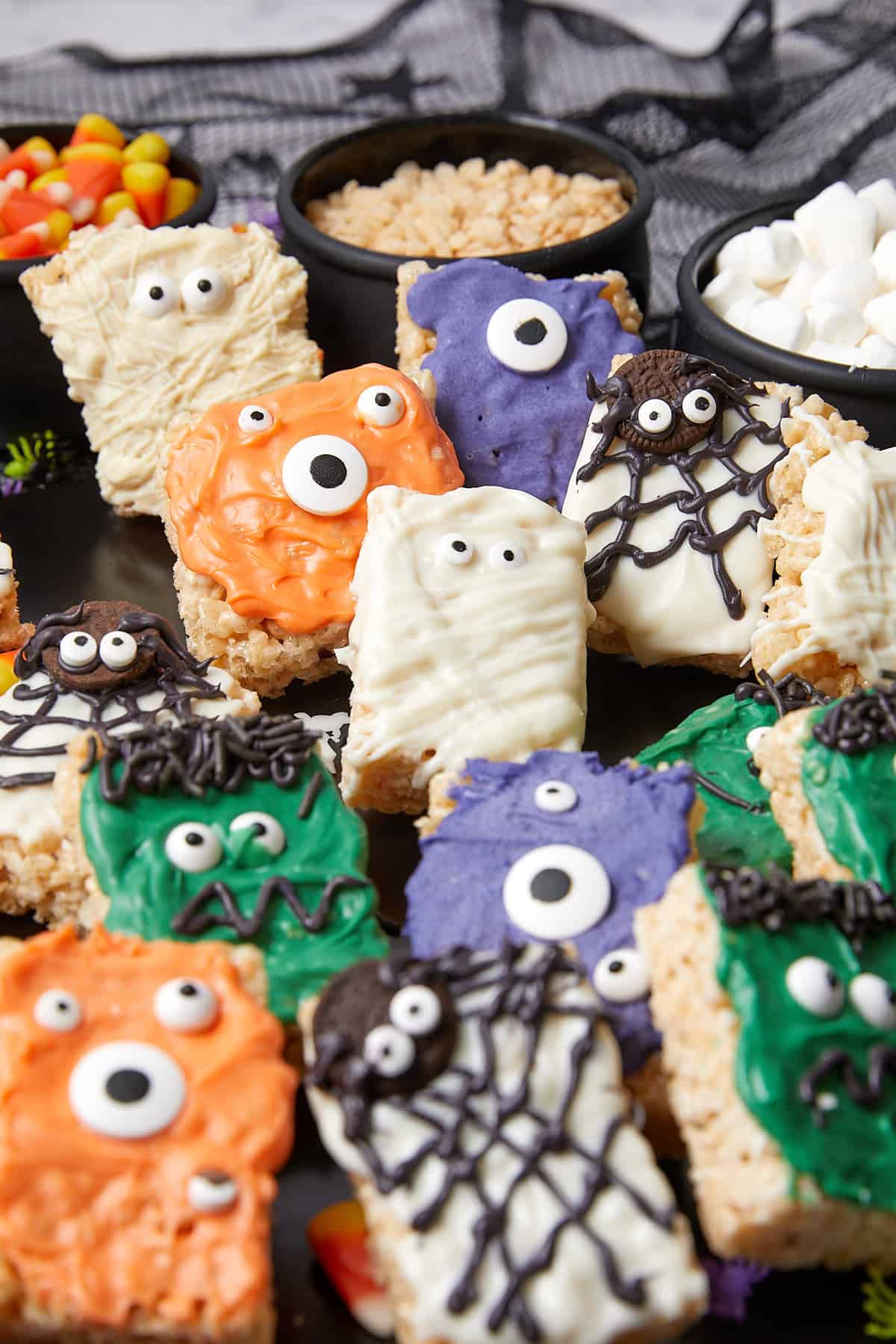A tray of Rice Krispies treats decorated as colorful Halloween monsters with googly eyes and spiderweb designs; bowls of candy corn, Rice Krispies, and mini marshmallows are in the background.