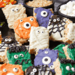 Colorful Rice Krispie treats decorated as Halloween monsters with candy eyes, icing in orange, white, purple, green, and black, and various spooky designs. Bowls of marshmallows, candy corn, and more treats are in the background.