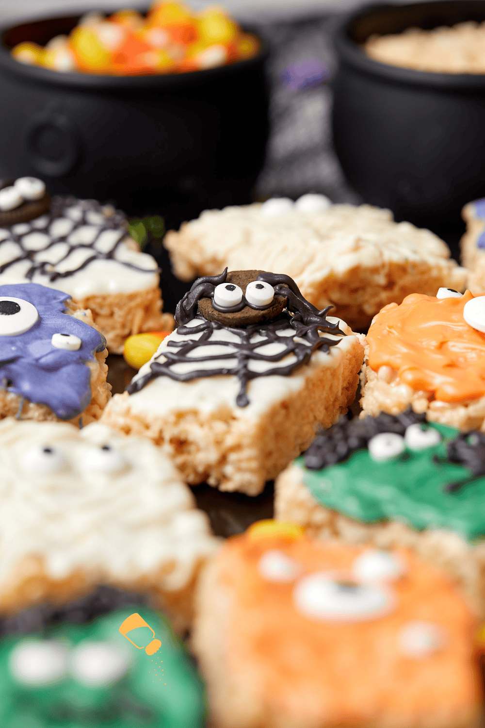 A close-up of Halloween-themed rice crispy treats decorated with colorful icing, candy eyes, and spooky designs like spiders and monsters. Black cauldrons and candy corn appear blurred in the background.