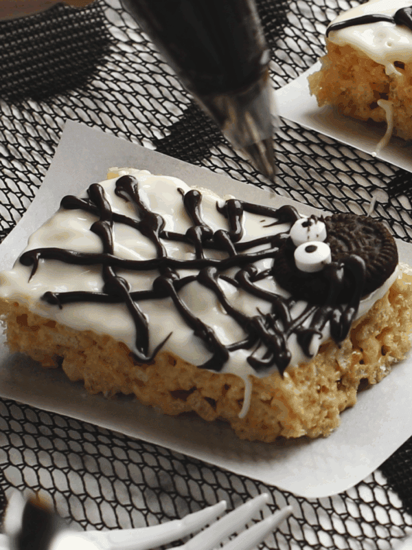 A hand decorates a rectangular Rice Krispies treat with white icing, chocolate web, and an Oreo cookie to resemble a spider on parchment paper.