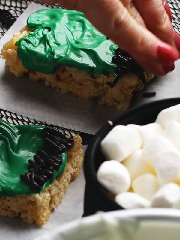 A hand decorates a green-frosted Rice Krispies treat shaped like Frankenstein’s face, next to a bowl of mini marshmallows.