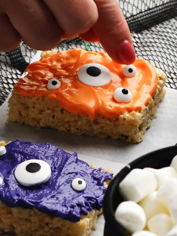 A hand adds a candy eyeball to an orange-frosted Rice Krispies treat decorated like a monster. Below it is a purple monster treat with candy eyes. A bowl of mini marshmallows sits in the corner.