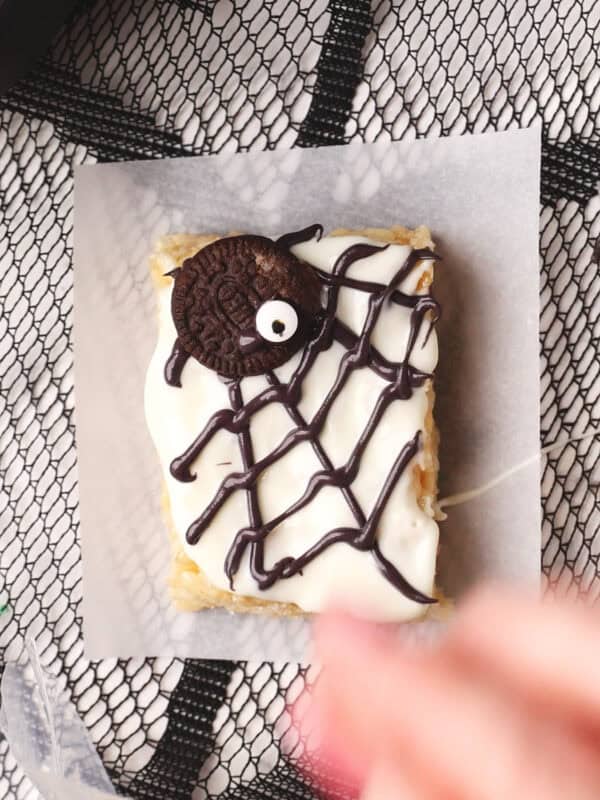A rectangular dessert topped with white icing, a chocolate spider made from a cookie and candy eye, and a chocolate web design, placed on parchment paper over a black mesh tablecloth.