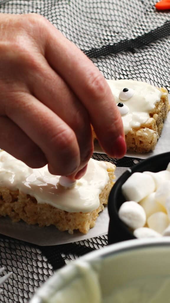 A hand places a candy eye on a frosted rice cereal treat shaped like a rectangle, with mini marshmallows in a bowl nearby and more decorated treats in the background.