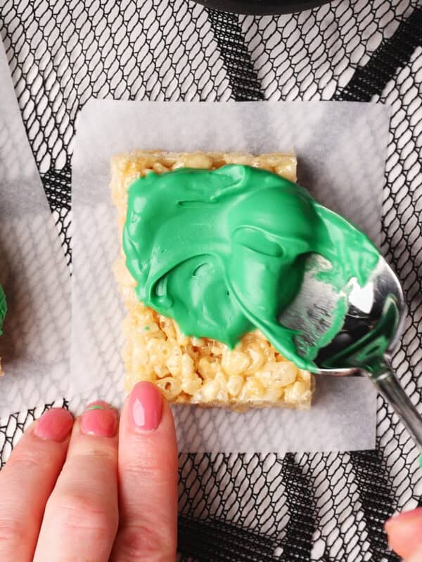 A hand spreads green icing onto a rectangular Rice Krispies treat using a spoon, with marshmallows visible in a bowl nearby on a textured black and white surface.