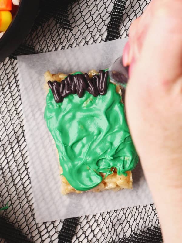 A hand decorates a rectangular rice crispy treat with green icing and black icing for hair on top, creating a Frankenstein monster look. The treat sits on parchment paper over a black mesh surface. Candy corn is visible in the corner.