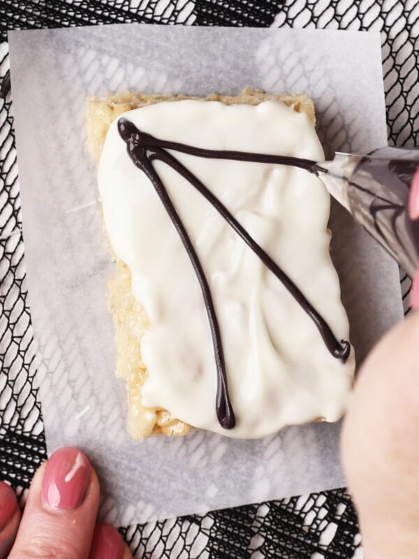 A hand pipes chocolate lines onto a rectangular rice krispie treat topped with white icing, placed on parchment paper over a black lace-patterned surface.