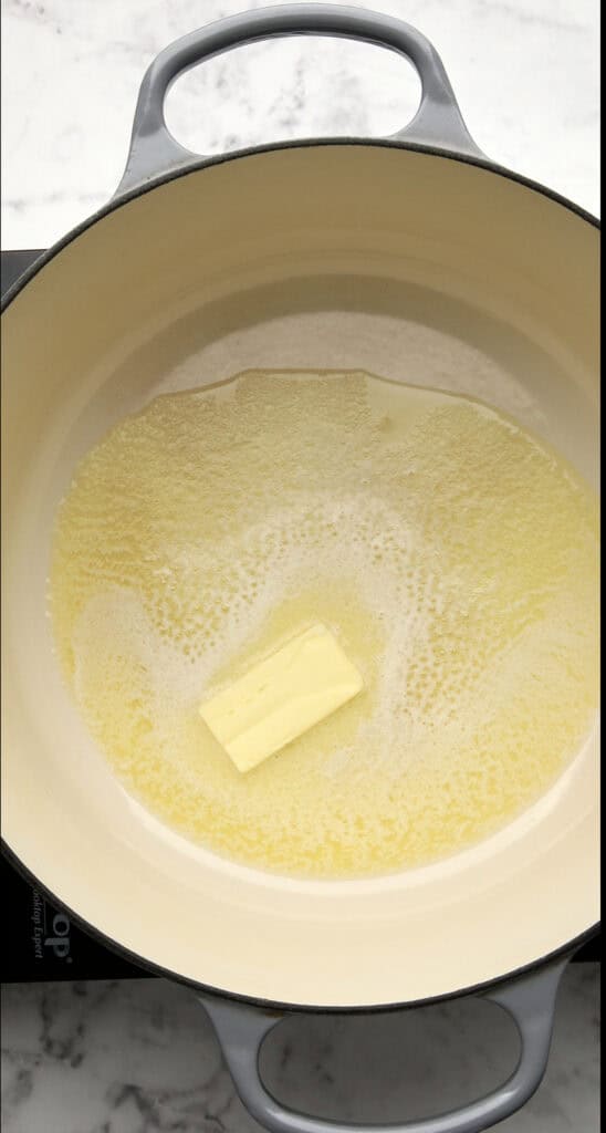 A half stick of butter melting in a large cream-colored pot on a stovetop with a marble countertop background.
