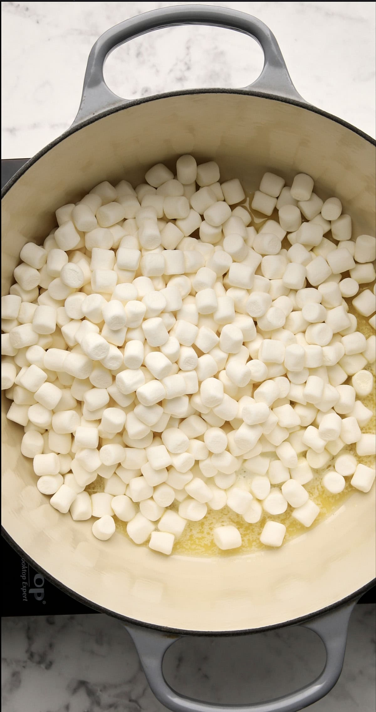 A large pot on a stovetop filled with mini marshmallows being melted over lightly melted butter, viewed from above on a marble countertop.