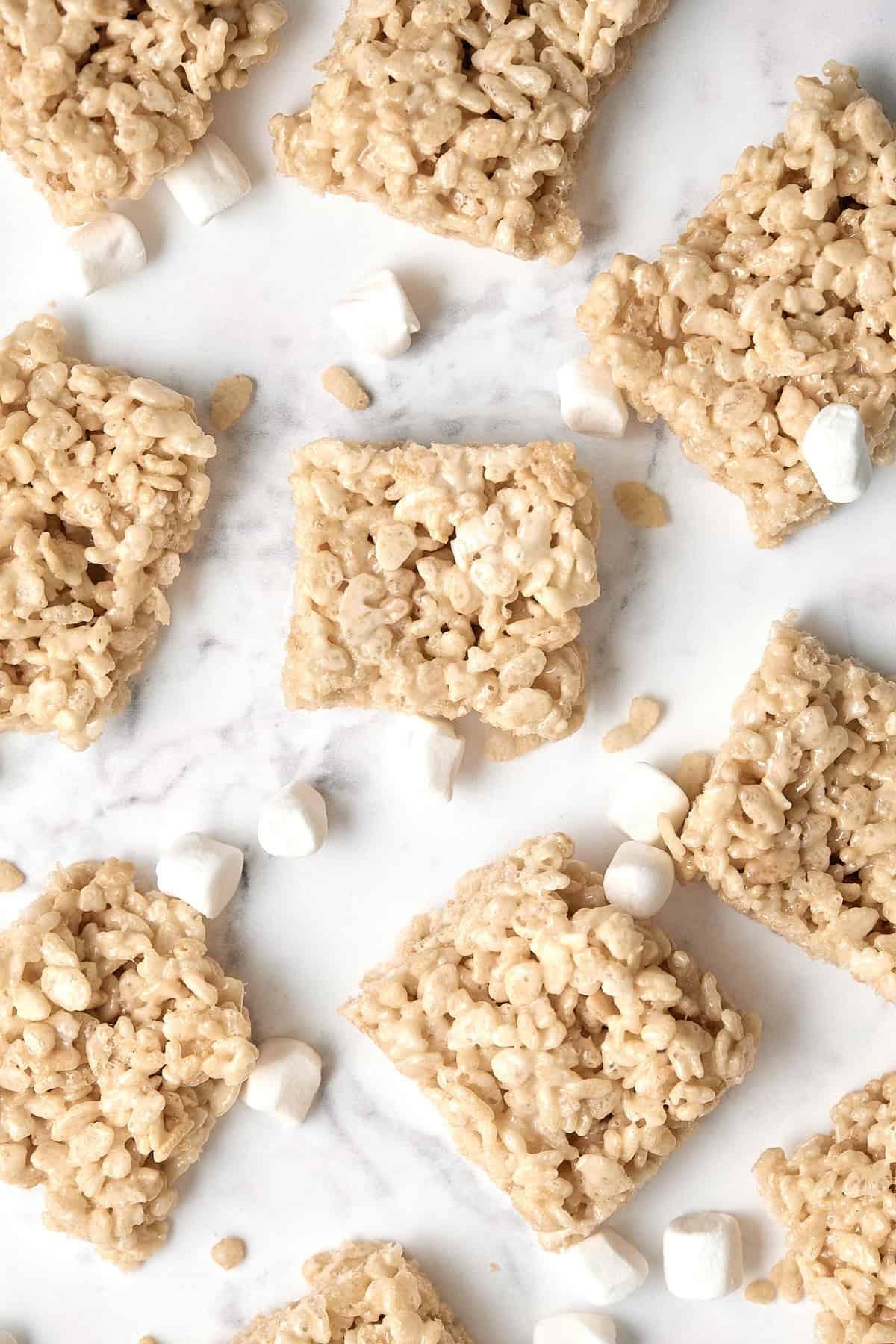 Squares of Rice Krispies treats are arranged on a white marble surface, surrounded by scattered mini marshmallows. The treats have a light, crispy texture and appear homemade.