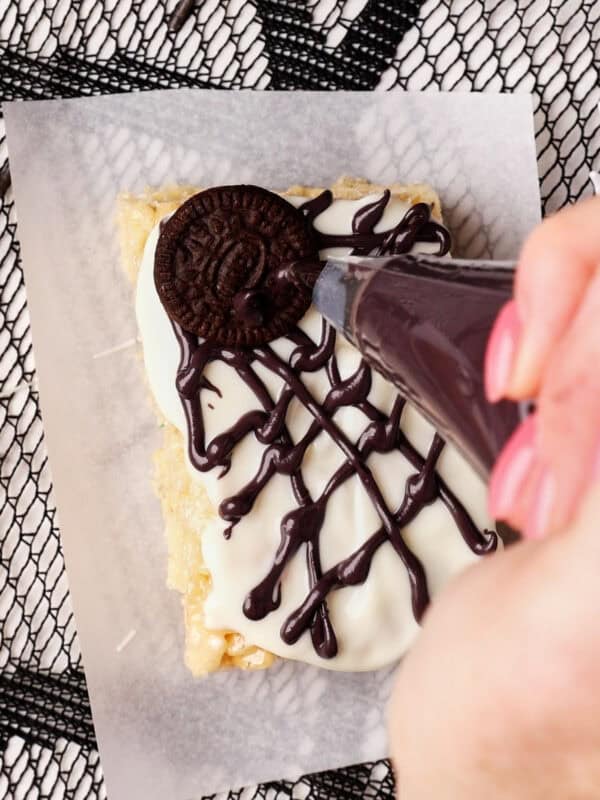 A hand decorates a rice crispy treat topped with white icing and a chocolate cookie by piping chocolate in a spiderweb pattern. The treat sits on parchment paper over a black mesh background.
