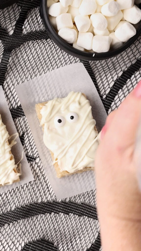 A rectangular treat covered in white icing with two candy eyes is being decorated to look like a mummy. It sits on parchment paper on a black spiderweb tablecloth, with a bowl of marshmallows nearby.