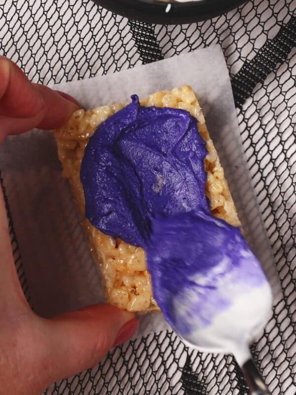 A hand spreads purple frosting onto a rice krispies treat with a spoon. A bowl of mini marshmallows is visible in the background on a black and white mesh surface.