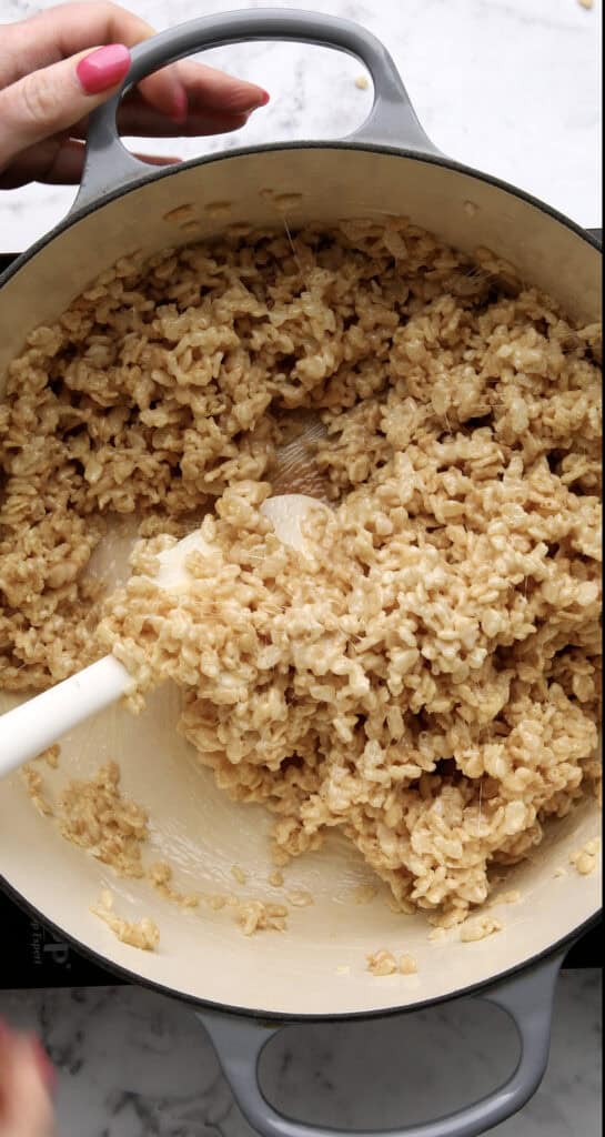 A hand holds a pot handle while another stirs gooey brown Rice Krispies treat mixture with a white spatula in a gray Dutch oven on a marble countertop.