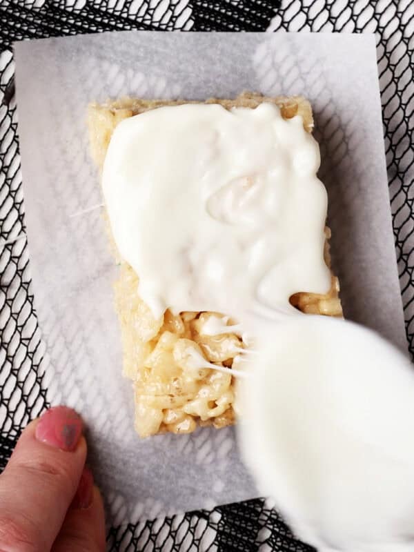 A hand holds a rice krispie treat on parchment paper while a spoon spreads white icing or frosting over the top. The background features a black mesh pattern.