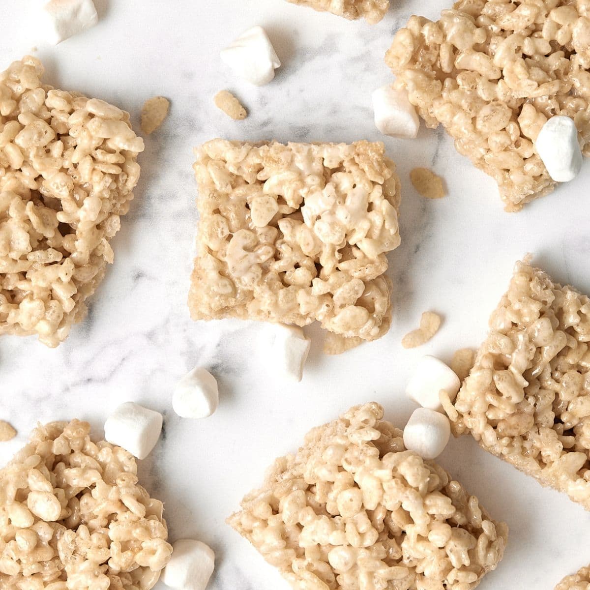 Rice Krispies treats and mini marshmallows are scattered on a white marble surface. The treats are square-shaped and made of puffed rice cereal and melted marshmallows.