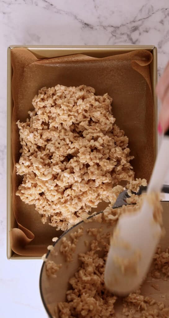 A person spreads a mixture of rice cereal and melted marshmallow into a parchment-lined square baking pan using a white spatula. The scene is viewed from above on a marble countertop.