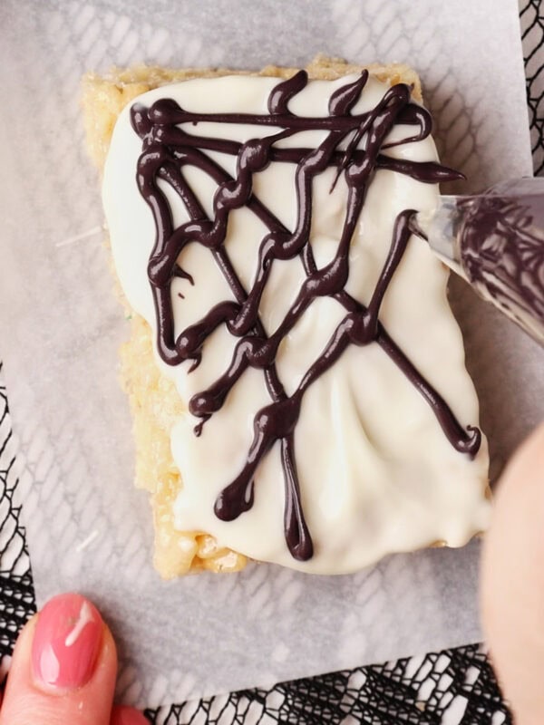 A hand decorates a frosted rectangular dessert with a chocolate spiderweb pattern using a piping bag, on a piece of parchment paper over a lace-patterned surface.