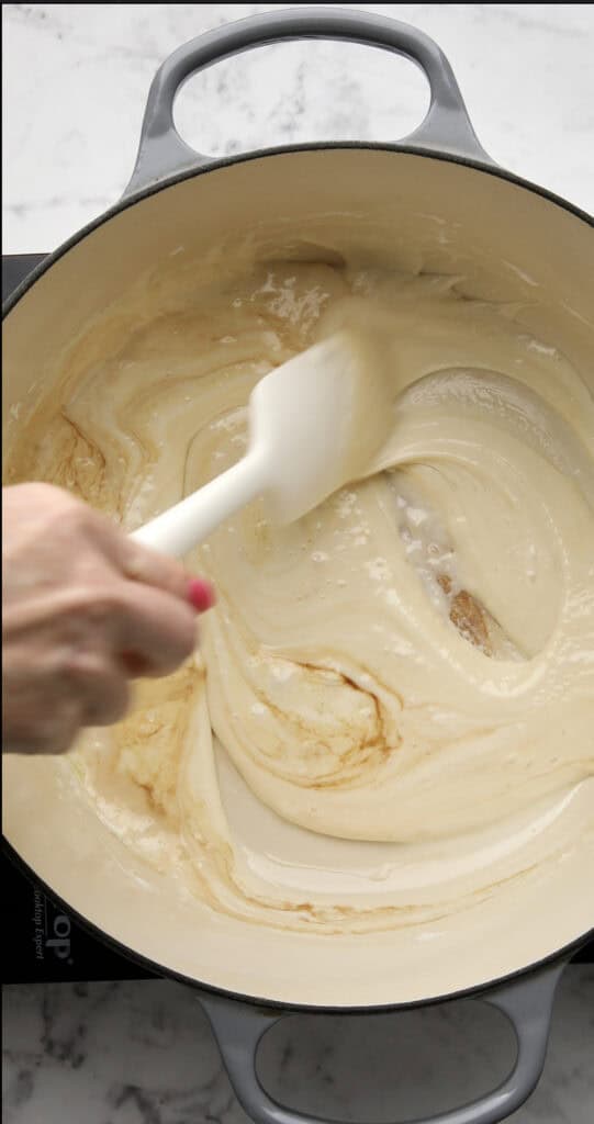 A hand holding a white spatula stirs a creamy, light brown sauce in a large, light-colored pot on a stovetop, with a marble countertop visible in the background.